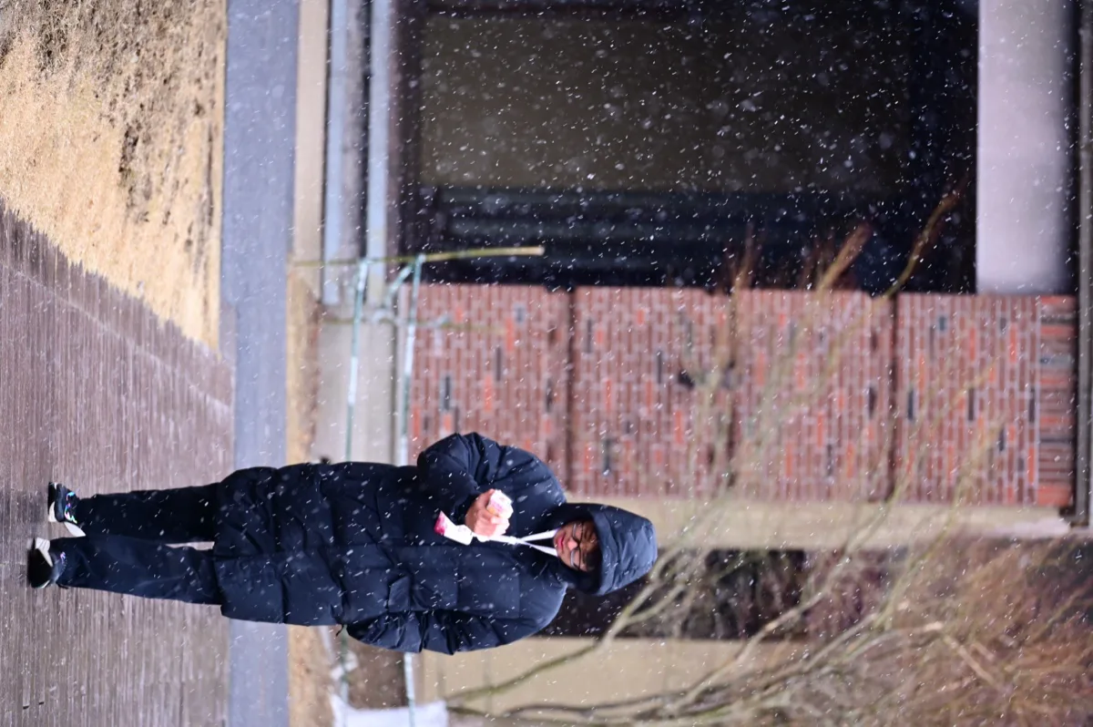 穿深色外套的人独自走过飞雪中的砖墙